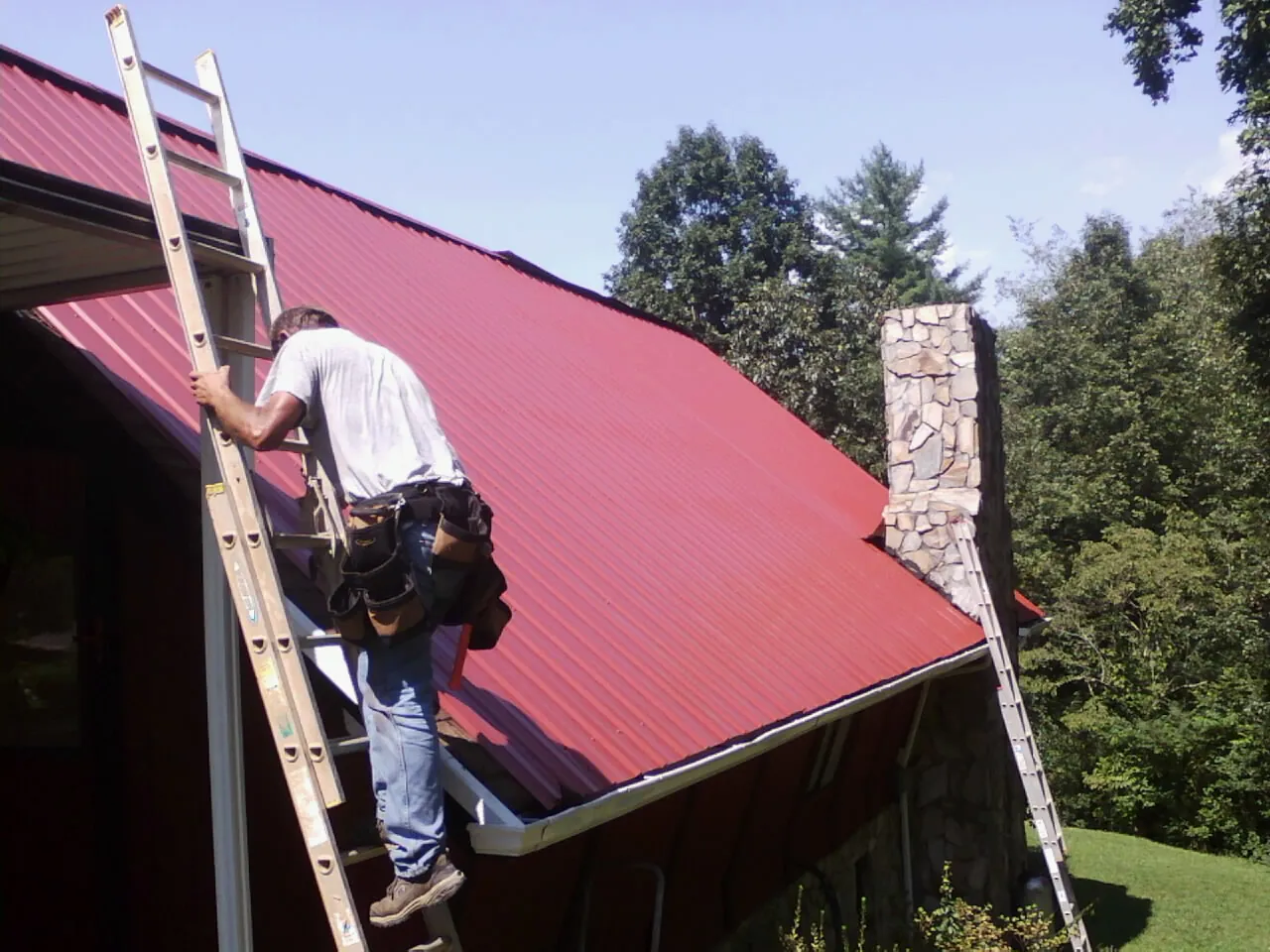 Roofer on a ladder beside a red metal roof and stone chimney — Soffit & Fascia Repair by Roofing Brothers in Flint Hill